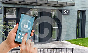 Female hand holding black mobile smart phone with smart home application on the screen. Blurred house on the background