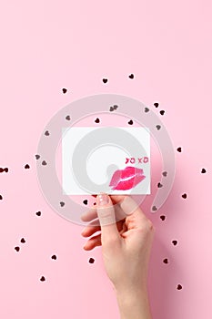 Female hand holding blank paper card mockup with lipstick kiss over pink background with confetti hearts. Happy Valentines Day or