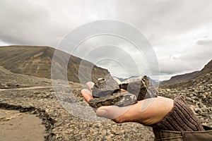 Female hand holding coal found by the Longyear glacier. Brown meltwater river and arctic landscape of glacial moraine