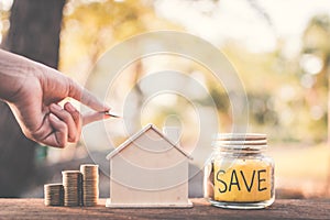 Female hand holding coins on glass jar with save word and model house on table in nature bokeh background