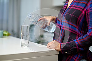 Female hand holding drinking water bottle and pouring water into glass on table on blurred kitchen background.