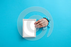 Female hand holding empty white wooden frame, body part sticking out of torn hole in blue paper background