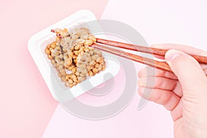 Female hand holding japanese natto beans with wooden chopsticks on pink background, top view.