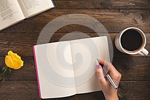 Female hand holding pen. Notebook, cup of coffee, rose, on wooden background