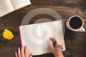 Female hand holding pen. Notebook, cup of coffee, rose, on wooden background.