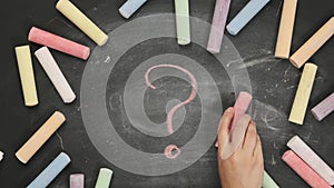 Female hand holding pink chalk and drawing a question mark on a black chalk board, top view, concept of answers and help