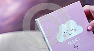 Female hand holding a pink diary with text DREAM BIG and nice cloudlet on a pink purple background. Inspiration and motivation photo