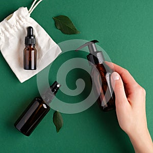 Female hand holding pump amber glass bottle over green background with spray bottle and essential oil dropper bottle. SPA natural
