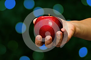 Female hand holding red apple on defocused abstract blue bokeh lights dark background. Close-up. Copy space