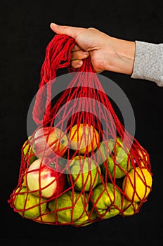 Female hand holding a red mesh bag with fruits on black background. Zero waste. Rejection of plastic bags concept