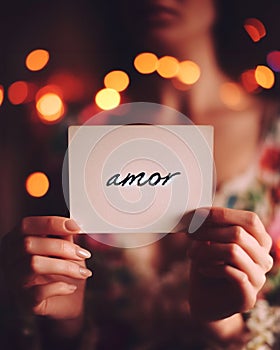 female hand holding a sign with spanish word amor or love , bokeh background lights, valentines concept