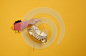 Female hand is holding a small golden clutch bag with a metal chain on a yellow background. Part of the body sticking out of a