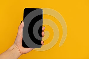 Female hand holding smartphone with black blank screen for mockup on yellow studio background with free space