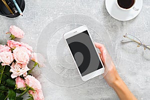 Female hand holding smartphone with blank screen mockup. Composition with roses flowers, coffee cup, stationery, glasses on