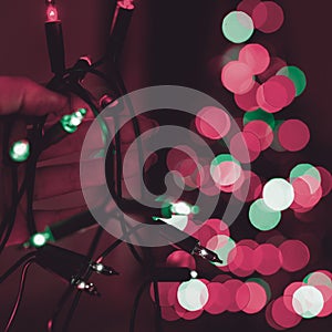 Female hand holding string of Christmas lights with defocused xmas tree in the background. Christmas lights.