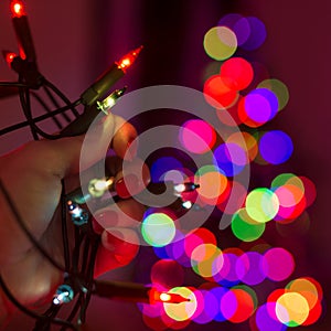 Female hand holding string of Christmas lights with defocused xmas tree in the background. Christmas lights bokeh.
