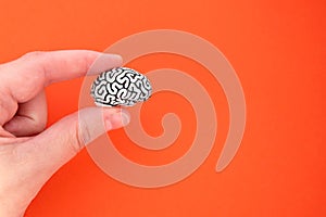 Female hand holding a tiny human brain model made of steel showing brain folds against an orange background