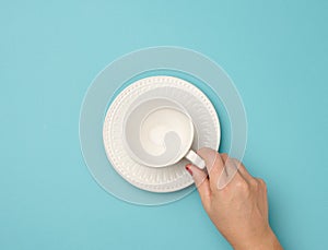 Female hand holding a white ceramic mug with a saucer on a blue background