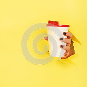 Female hand holding white paper mug on yellow background. Take away coffee cup concept. Mock up with copy space. Square crop