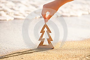 Female hand holding wooden Christmas tree on a sand on a beach near ocean, summer Christmas and winter holyday concept