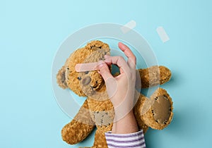 Female hand holds a brown teddy bear and glues a medical adhesive plaster on a blue background, tram treatment