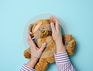 Female hand holds a brown teddy bear and glues a medical adhesive plaster on a blue background, tram treatment