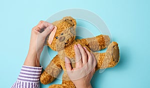 Female hand holds a brown teddy bear and glues a medical adhesive plaster on a blue background, tram treatment
