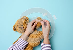 Female hand holds a brown teddy bear and glues a medical adhesive plaster on a blue background, tram treatment