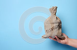 A female hand holds a burlap sack with the inscription subsidy on a blue background, concept of increasing government subsidies to