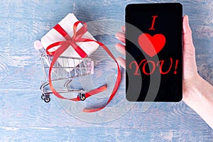 A female hand holds a cellphone with the inscription I LOVE YOU and a shopping trolley with a gift box