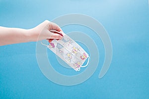 female hand holds children`s medical protective mask with drawings on a blue background copy space, hand without gloves, protecti