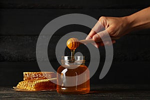 Female hand holds dipper honey on wooden background with jar and honeycombs