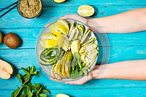 Female hand holds fresh salad with avocado, kiwi, apple, cucumber, pear, lime and mint with smoothie on blue wooden