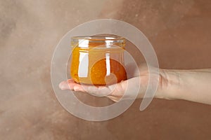 Female hand holds glass jar apricot jam on brown background