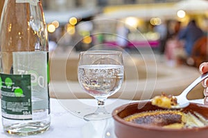 A female hand holds a large spoon of creme brulee dessert with a glass and bottle of sparkling water at an outdoor cafe