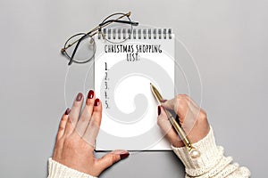 Female hand holds pen, writing Christmas shopping list, gift ideas on white notepad on gray background Top view Flat lay Holiday