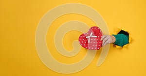 Female hand holds a red box with a gift on a yellow background, part of the body sticks out of a torn hole in a paper background.