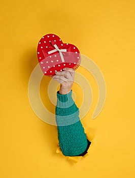 Female hand holds a red box with a gift on a yellow background, part of the body sticks out of a torn hole in a paper background.