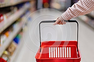 Female hand holds and shows empty red grocery cart, close-up. In background is shopping mall with shelves in store