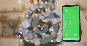 Female hand holds smartphone with green screen, flashing garland on Christmas tree on the background. Close-up, space