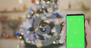 Female hand holds smartphone with green screen, flashing garland on Christmas tree on the background. Close-up, space