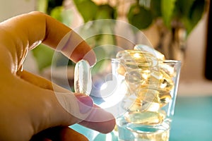 female hand holds a transparent omega 3 capsule, a glass with tablets on a table near the branches of a birch .