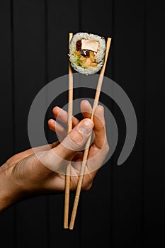 female hand holds unagi sushi roll with smoked eel with chopsticks on dark background