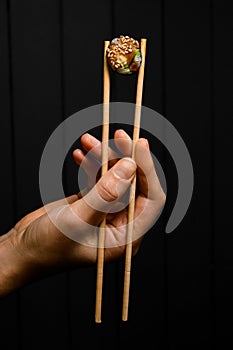 female hand holds vegetarian sushi roll with bamboo chopsticks on dark background