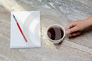 Female hand holds a white cup of coffee stands on the table. Notepad and fountain pen