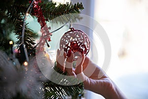 Female hand with holiday manicure holding shiny red holiday bauble hanging from a christmas tree