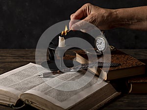 A female hand lights a candle on a table with old books, a quill pen and an antique pocket watch