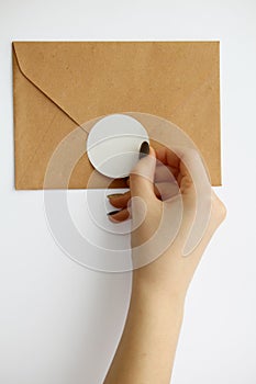 female hand with manicure glues a round sticker on a brown envelope on a white background.  mockup stickers with plotter cutting