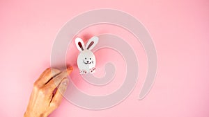 Female hand near a cute Easter bunny made from a white chicken egg isolated on a light pink pastel background
