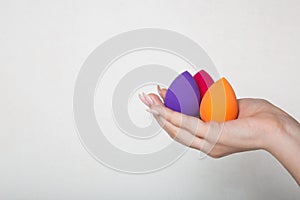 Female hand with nice manicure holding purple, pink and orange beauty blenders. Space for text